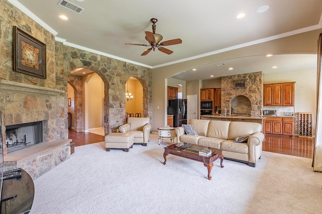 living room with arched walkways, a stone fireplace, crown molding, a ceiling fan, and light colored carpet