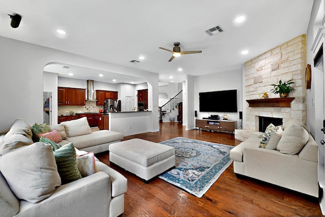 living room with ceiling fan, arched walkways, dark wood finished floors, a stone fireplace, and stairway