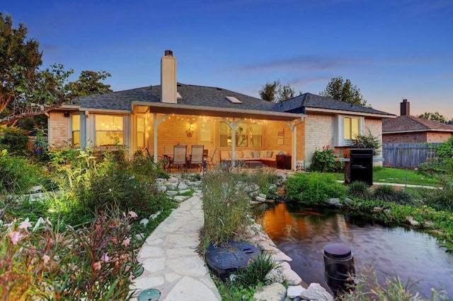 back of property at dusk featuring fence, a chimney, brick siding, a patio, and a garden pond