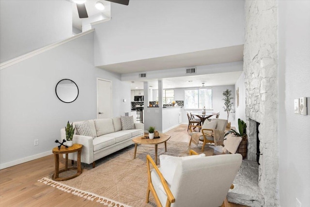 living room featuring light wood finished floors, a stone fireplace, a ceiling fan, baseboards, and a towering ceiling
