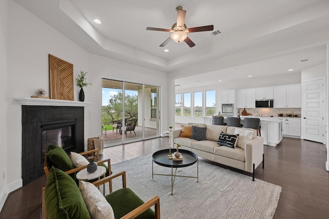 living room with a glass covered fireplace, a tray ceiling, recessed lighting, a ceiling fan, and dark wood-style flooring