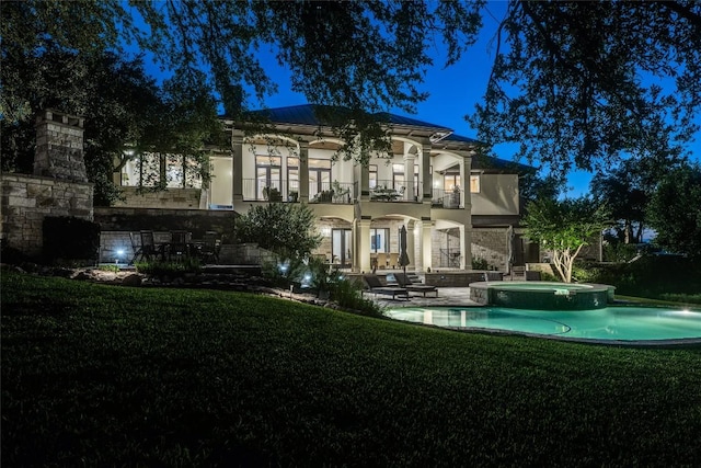 back of house at night featuring a balcony, a lawn, a patio area, and stucco siding
