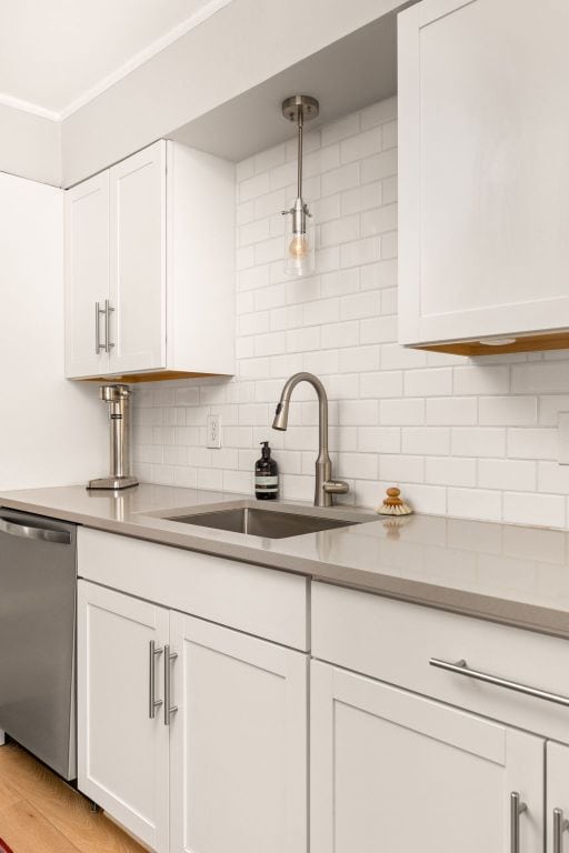 kitchen featuring stainless steel dishwasher, a sink, white cabinets, backsplash, and hanging light fixtures