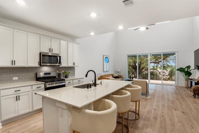 kitchen with appliances with stainless steel finishes, a sink, light wood-type flooring, a kitchen island with sink, and decorative backsplash