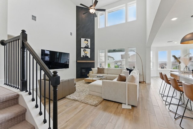 living room featuring a towering ceiling, healthy amount of natural light, light wood-type flooring, ceiling fan, and a fireplace