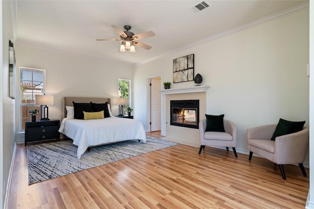 bedroom with light wood-style flooring, crown molding, a glass covered fireplace, ceiling fan, and baseboards