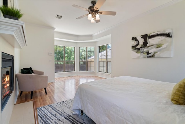 bedroom featuring ornamental molding, a glass covered fireplace, light wood finished floors, and ceiling fan
