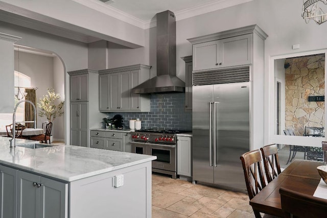 kitchen with high end appliances, gray cabinets, wall chimney range hood, light stone counters, and decorative backsplash