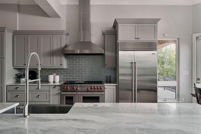 kitchen featuring high quality appliances, gray cabinets, wall chimney range hood, decorative backsplash, and light stone counters