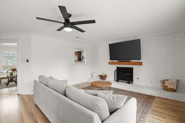 living area with a brick fireplace, light wood-style flooring, ceiling fan, crown molding, and brick wall