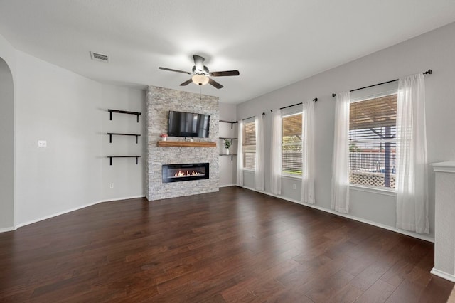unfurnished living room with arched walkways, a stone fireplace, a ceiling fan, dark wood-style floors, and baseboards