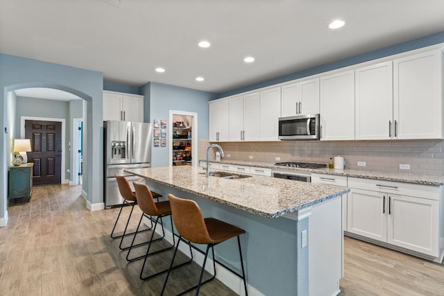 kitchen featuring stainless steel appliances, a sink, arched walkways, a kitchen island with sink, and decorative backsplash
