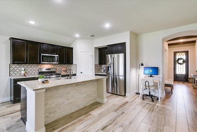 kitchen with stainless steel appliances, arched walkways, a sink, light wood-type flooring, and an island with sink