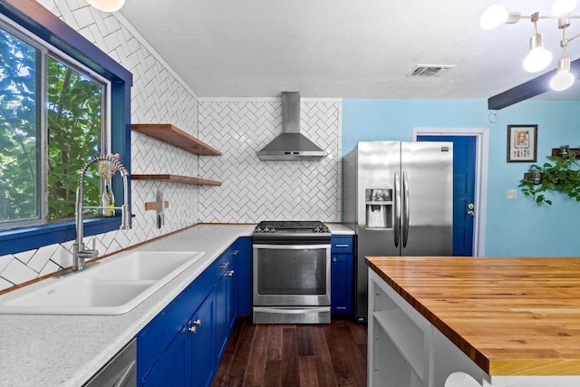 5105 Stone Gate Dr, Austin, TX 78721 - kitchen with stainless steel appliances, wall chimney exhaust hood, blue cabinets, wooden counters, and decorative backsplash