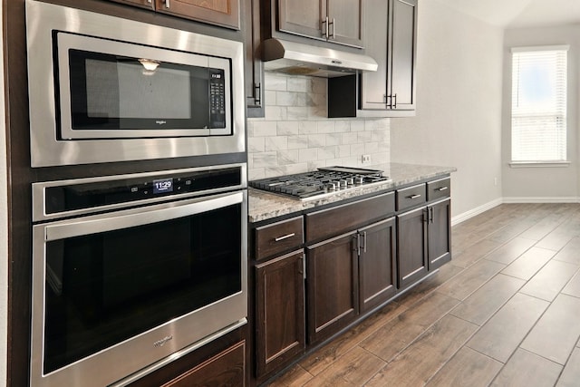 kitchen with appliances with stainless steel finishes, under cabinet range hood, baseboards, decorative backsplash, and dark wood-style flooring