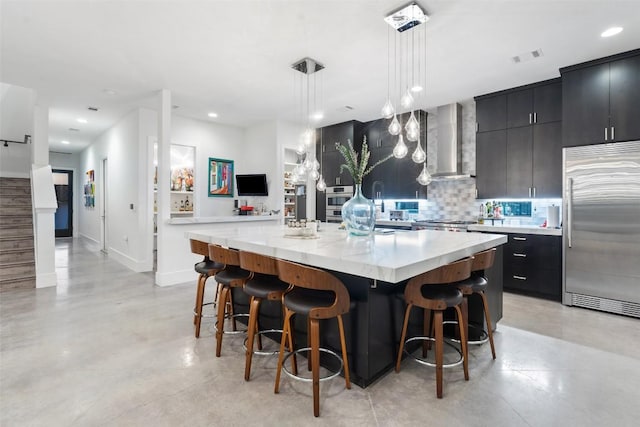 kitchen with stainless steel built in refrigerator, wall chimney range hood, dark cabinets, decorative backsplash, and recessed lighting