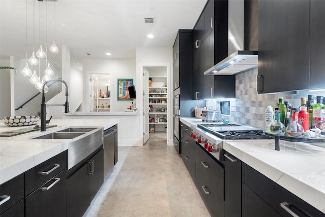 kitchen featuring wall chimney exhaust hood, dark cabinetry, stainless steel appliances, a sink, and hanging light fixtures
