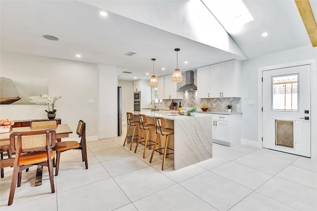 kitchen featuring wall chimney range hood, backsplash, white cabinets, an island with sink, and recessed lighting