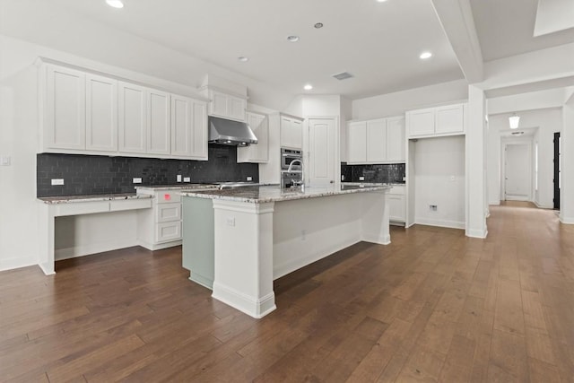 kitchen with under cabinet range hood, decorative backsplash, white cabinets, an island with sink, and dark wood-type flooring