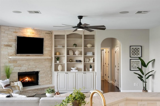 living room with arched walkways, a stone fireplace, a ceiling fan, baseboards, and dark wood finished floors