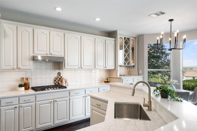 kitchen featuring stainless steel gas cooktop, a sink, under cabinet range hood, a chandelier, and white cabinetry