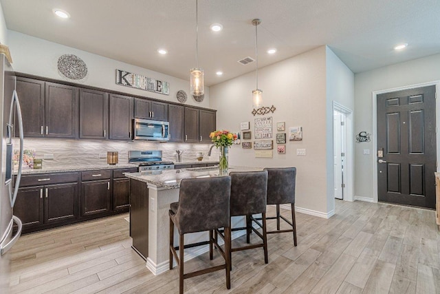 kitchen featuring appliances with stainless steel finishes, light wood finished floors, dark brown cabinets, light stone countertops, and a kitchen bar