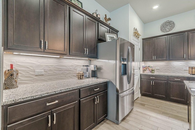 kitchen with stainless steel fridge, decorative backsplash, light wood-type flooring, dark brown cabinets, and light stone countertops