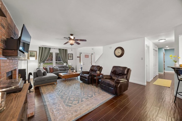 living area with a brick fireplace, a ceiling fan, dark wood-type flooring, and baseboards