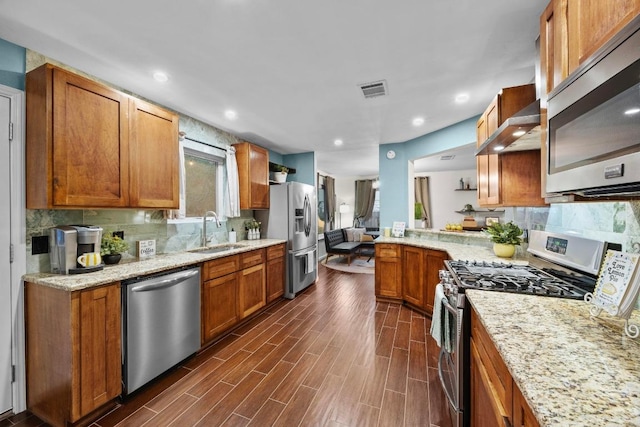 kitchen with appliances with stainless steel finishes, a sink, wall chimney exhaust hood, brown cabinetry, and backsplash