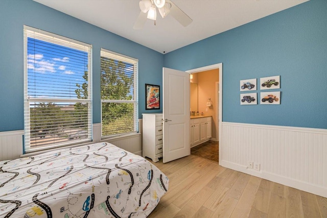 bedroom featuring a wainscoted wall, light wood-type flooring, ceiling fan, and ensuite bath