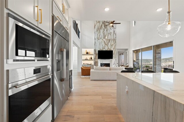 6901 Davenport Divide Rd, Austin, TX 78738 - kitchen featuring appliances with stainless steel finishes, a towering ceiling, light wood-style flooring, light stone counters, and a fireplace
