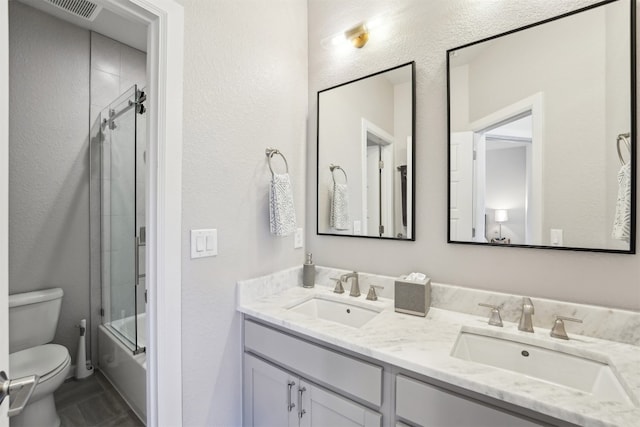 full bath featuring toilet, double vanity, a textured wall, and shower / bath combination with glass door