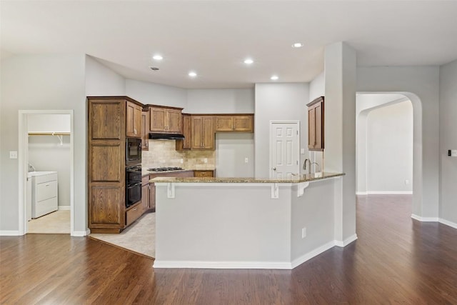 5501 Wild Foxglove Rd, Spicewood, TX 78669 - kitchen with washer / dryer, black microwave, a peninsula, brown cabinetry, and light stone counters