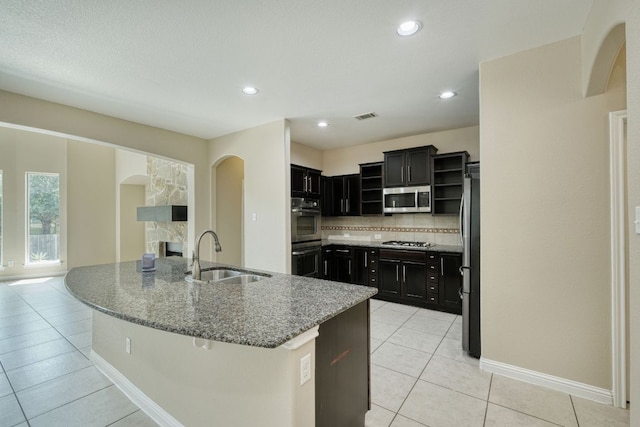 2937 Castellan Ln, Round Rock, TX 78665 - kitchen with appliances with stainless steel finishes, open shelves, an island with sink, light tile patterned floors, and dark stone countertops
