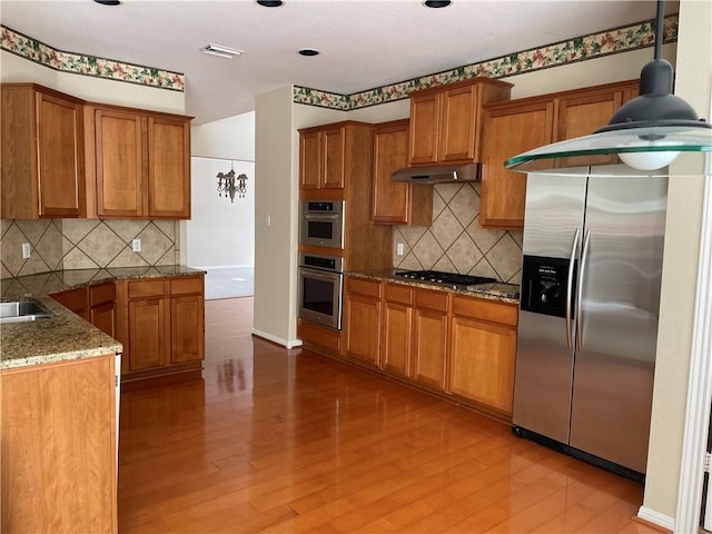 kitchen featuring stainless steel appliances, under cabinet range hood, brown cabinetry, and backsplash