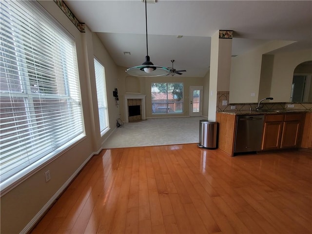 kitchen featuring stainless steel dishwasher, brown cabinets, a ceiling fan, a tiled fireplace, and light wood-style floors