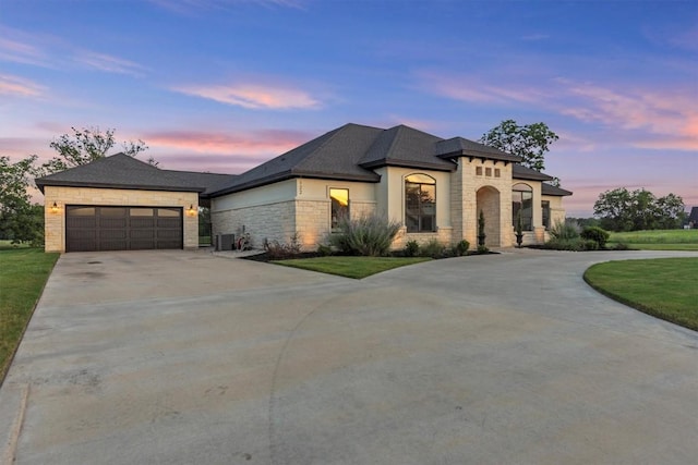 french country home featuring concrete driveway, stone siding, central AC, an attached garage, and a yard