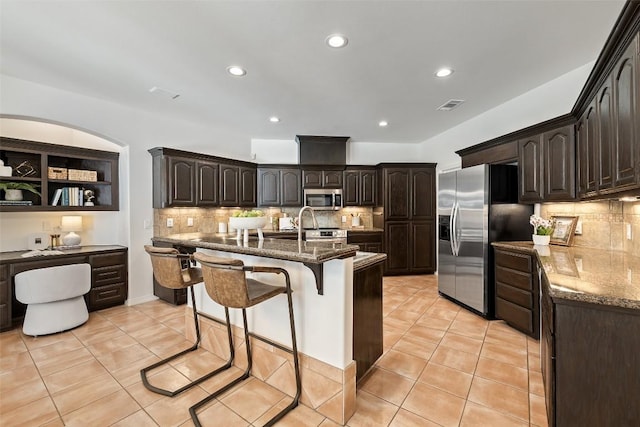 8311 Sumner Ct, Austin, TX 78733 - kitchen featuring appliances with stainless steel finishes, light tile patterned flooring, dark stone counters, a kitchen breakfast bar, and tasteful backsplash