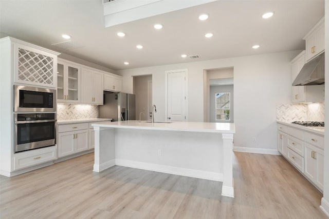 159 Fireside Rd, Austin, TX 78737 - kitchen featuring appliances with stainless steel finishes, under cabinet range hood, light wood finished floors, an island with sink, and tasteful backsplash