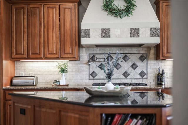 17400 Northlake Hills Dr, Jonestown, TX 78645 - kitchen with wall chimney exhaust hood, tasteful backsplash, brown cabinetry, stainless steel gas stovetop, and dark countertops