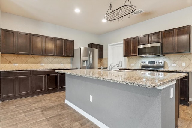 1008 Comal Run, Hutto, TX 78634 - kitchen featuring appliances with stainless steel finishes, a sink, dark brown cabinets, a kitchen island with sink, and light wood finished floors