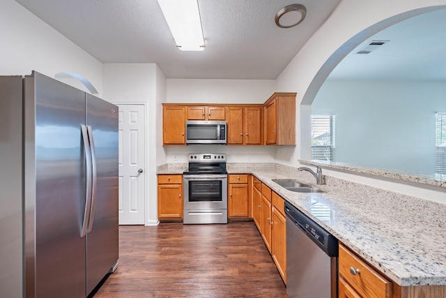 18111 Gallant St, Manor, TX 78653 - kitchen featuring appliances with stainless steel finishes, arched walkways, dark wood-style floors, light stone countertops, and brown cabinets