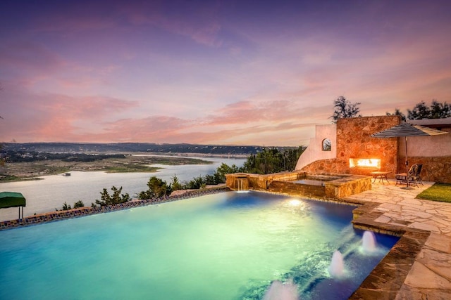 908 Watercliffe Dr, Lago Vista, TX 78645 - pool at dusk with an outdoor stone fireplace, a patio, a water view, and a pool with connected hot tub
