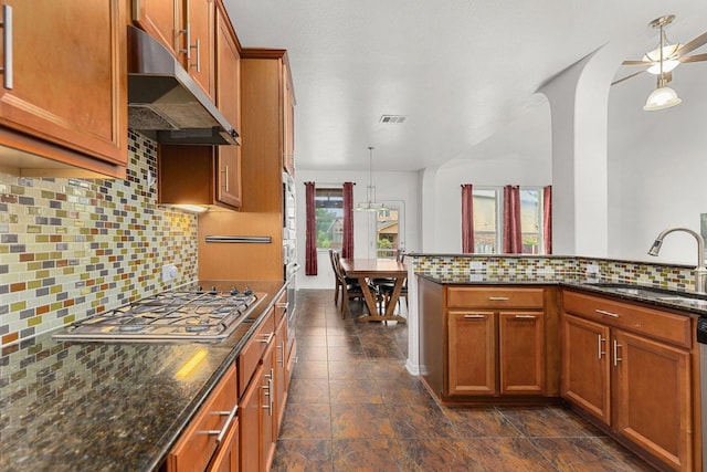 204 Lake Livingston Dr, Georgetown, TX 78628 - kitchen featuring stainless steel gas stovetop, under cabinet range hood, backsplash, brown cabinets, and hanging light fixtures