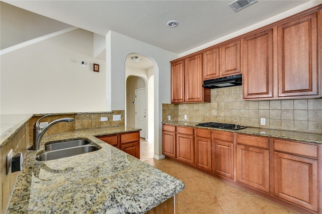 903 Fork Ridge Path, Round Rock, TX 78665 - kitchen featuring under cabinet range hood, light stone counters, tasteful backsplash, arched walkways, and brown cabinetry