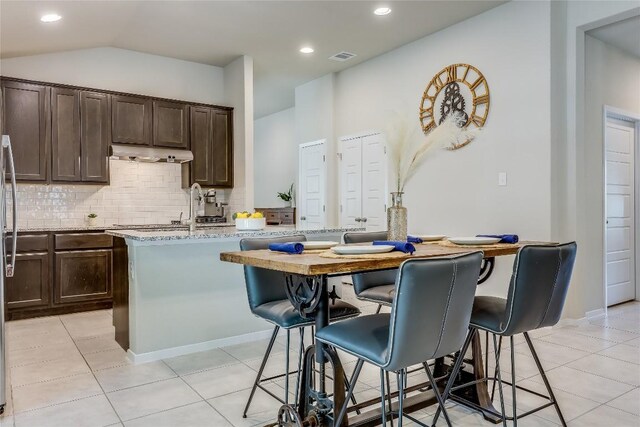 1901 Owens Ln, Lago Vista, TX 78645 - kitchen featuring under cabinet range hood, dark brown cabinetry, decorative backsplash, a kitchen breakfast bar, and light stone counters