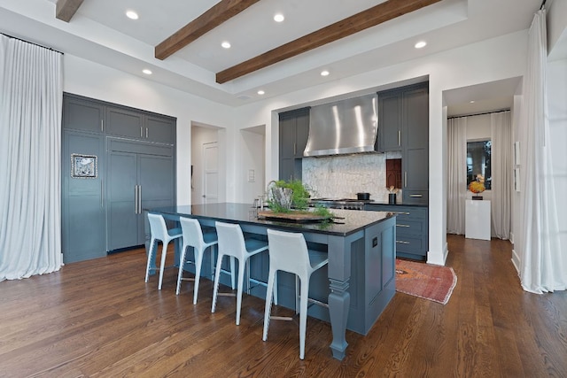 4716 Peralta Ln, Austin, TX 78735 - kitchen featuring wall chimney range hood, dark countertops, tasteful backsplash, beamed ceiling, and a kitchen island with sink