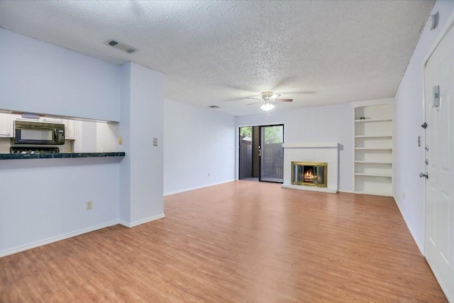 1202 Thorpe Ln Unit 407, San Marcos, TX 78666 - unfurnished living room featuring a textured ceiling, a ceiling fan, a brick fireplace, light wood-style flooring, and built in shelves
