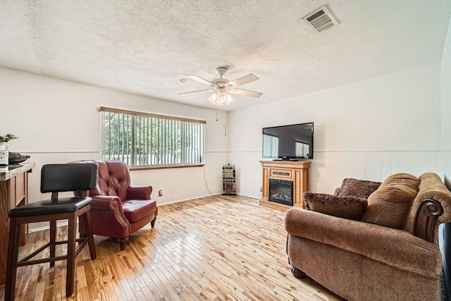 914 Hermitage Dr, Austin, TX 78753 - living room featuring a wainscoted wall, light wood finished floors, a textured ceiling, a ceiling fan, and a glass covered fireplace