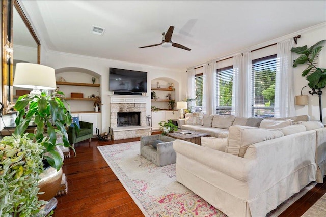 living area with ceiling fan, dark wood-type flooring, crown molding, a stone fireplace, and built in features